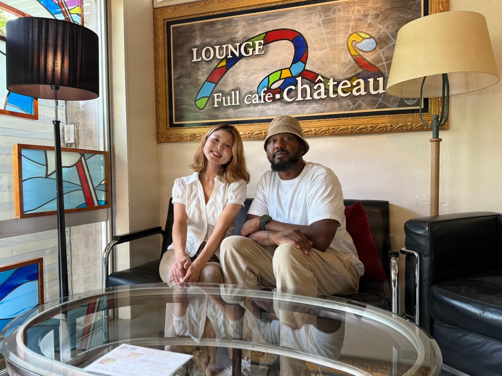 Two people are seated together on a black leather sofa inside a lounge cafe called 'Lounge Full Cafe Château,' posing for a photo. The interior features eclectic decor including stained glass panels, floor lamps, and a glass coffee table in the foreground. The atmosphere appears casual and relaxed.
