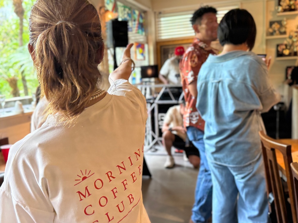 An indoor gathering or event where people are socializing near what appears to be a DJ setup. The foreground features a person wearing a white long-sleeve shirt with 'Morning Coffee Club' printed on the back along with a sun graphic. The setting appears to be a casual, home-like or café venue with natural light coming through windows.