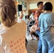 An indoor gathering or event where people are socializing near what appears to be a DJ setup. The foreground features a person wearing a white long-sleeve shirt with 'Morning Coffee Club' printed on the back along with a sun graphic. The setting appears to be a casual, home-like or café venue with natural light coming through windows.