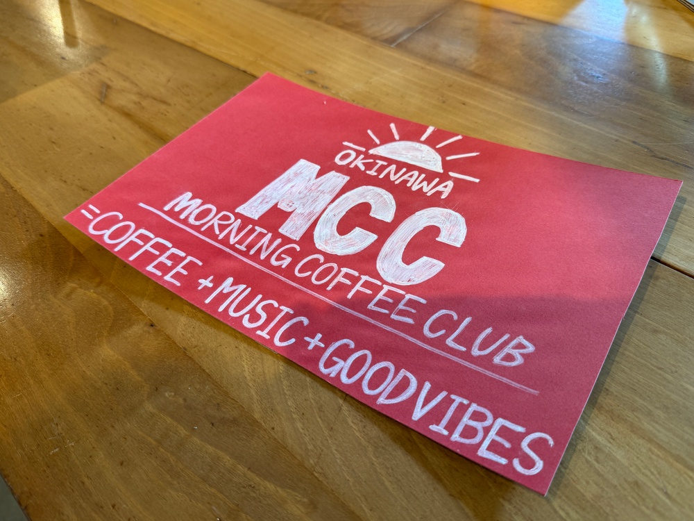 A red promotional flyer or sign for the 'Okinawa MCC Morning Coffee Club' is lying flat on a wooden table. The sign is hand-lettered in white chalk-style writing and features a sunrise logo above the acronym 'MCC'. It promotes a social gathering centered around coffee, music, and good vibes.