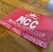 A red promotional flyer or sign for the 'Okinawa MCC Morning Coffee Club' is lying flat on a wooden table. The sign is hand-lettered in white chalk-style writing and features a sunrise logo above the acronym 'MCC'. It promotes a social gathering centered around coffee, music, and good vibes.