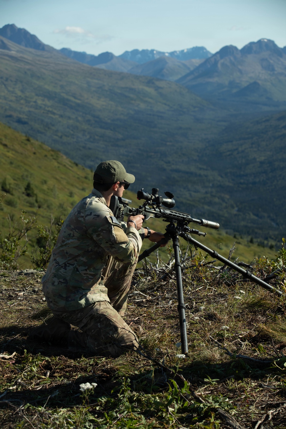 A person in military-style multicam camouflage uniform is kneeling on a hillside, aiming a precision rifle mounted on a tripod. The setting appears to be a mountainous, heavily forested region with snow-capped peaks visible in the background under clear skies. The scene suggests a long-range shooting exercise or training activity at elevation.
