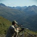 A person in military-style multicam camouflage uniform is kneeling on a hillside, aiming a precision rifle mounted on a tripod. The setting appears to be a mountainous, heavily forested region with snow-capped peaks visible in the background under clear skies. The scene suggests a long-range shooting exercise or training activity at elevation.