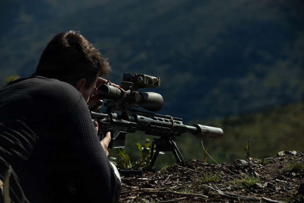 A person is lying in a prone position on rocky, outdoor terrain while aiming a scoped precision rifle. The rifle is mounted on a bipod and equipped with multiple optics including a magnified scope and a secondary sighting device. The background shows a blurred mountainous or hilly landscape.
