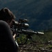 A person is lying in a prone position on rocky, outdoor terrain while aiming a scoped precision rifle. The rifle is mounted on a bipod and equipped with multiple optics including a magnified scope and a secondary sighting device. The background shows a blurred mountainous or hilly landscape.