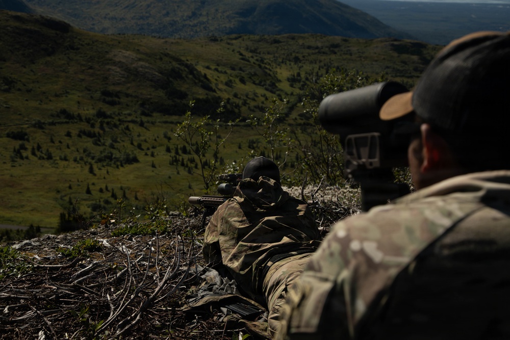 Two military personnel in camouflage uniforms are positioned on an elevated ridge overlooking a vast green valley. One individual is in a prone shooting position with a scoped rifle, while the second is using a large spotting scope. The setting appears to be a highland or mountainous training environment during daylight hours.