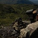 Two military personnel in camouflage uniforms are positioned on an elevated ridge overlooking a vast green valley. One individual is in a prone shooting position with a scoped rifle, while the second is using a large spotting scope. The setting appears to be a highland or mountainous training environment during daylight hours.