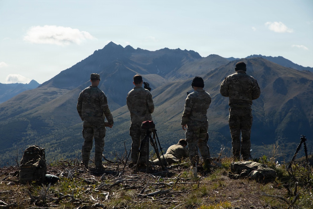 Four military personnel in camouflage uniforms stand on an elevated ridge overlooking a dramatic mountain range, likely during a training exercise. They appear to be conducting observation or reconnaissance activities, with one individual crouched near equipment on the ground. The setting suggests a mountainous region, possibly Alaska or a similar high-altitude terrain.