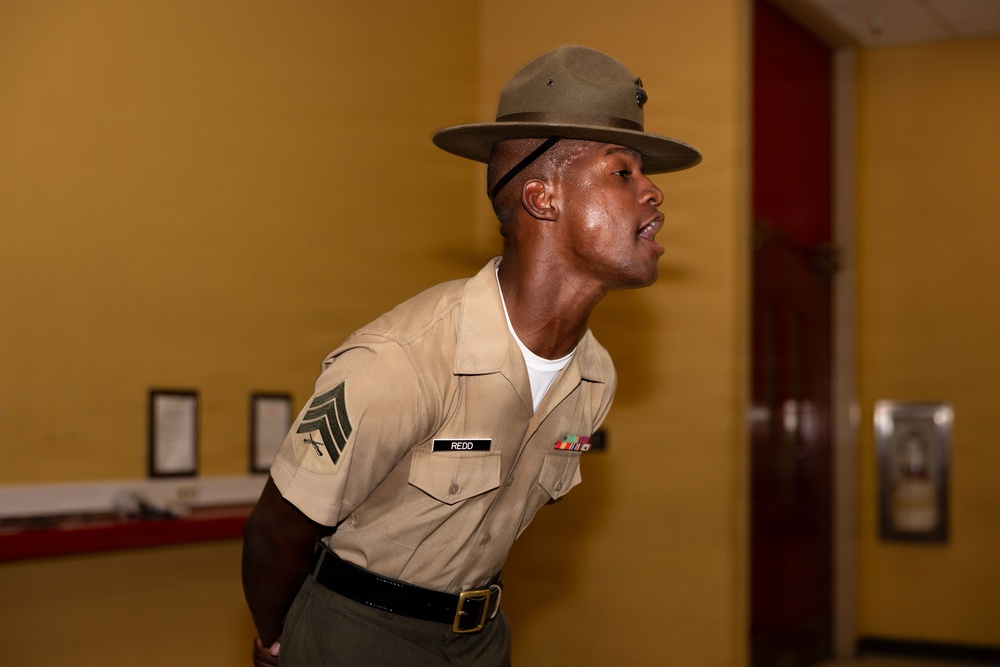 A U.S. Marine Corps Drill Instructor in service uniform is photographed in a leaning forward posture, appearing to be issuing commands or instructions. The Marine wears the distinctive Drill Instructor campaign cover (cover/hat) and displays the rank of Staff Sergeant on the left sleeve. The name tape reads 'REDD' and several service ribbons are visible on the chest.