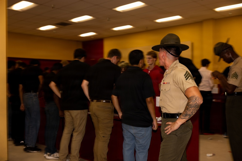 A U.S. Marine Corps staff sergeant or drill instructor, wearing the distinctive campaign cover (drill instructor hat) and tan service uniform with ribbons, oversees a group of civilians in an indoor setting. The civilians, mostly wearing dark clothing, appear to be recruits or potential enlistees at what looks like a Military Entrance Processing Station (MEPS) or recruitment event. A second uniformed Marine is visible on the right side of the frame.