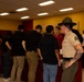A U.S. Marine Corps staff sergeant or drill instructor, wearing the distinctive campaign cover (drill instructor hat) and tan service uniform with ribbons, oversees a group of civilians in an indoor setting. The civilians, mostly wearing dark clothing, appear to be recruits or potential enlistees at what looks like a Military Entrance Processing Station (MEPS) or recruitment event. A second uniformed Marine is visible on the right side of the frame.