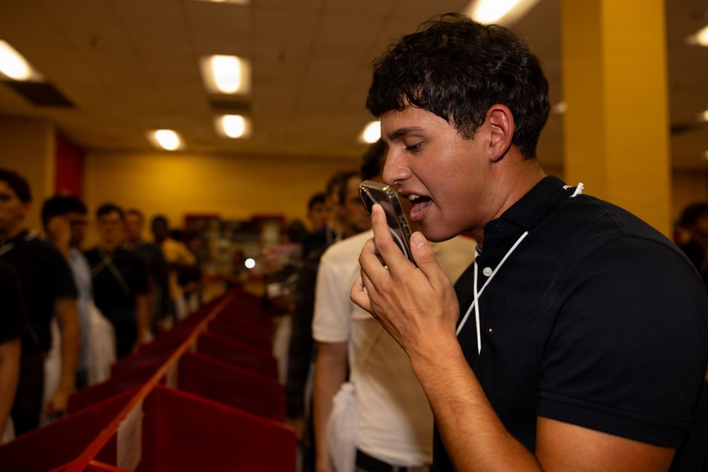 A young man in a black polo shirt is speaking into or recording with a smartphone held close to his mouth in what appears to be a retail store. A crowd of mostly young males is visible in the blurred background, suggesting a group event or gathering. The foreground subject is sharply in focus while the background is intentionally blurred.