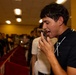 A young man in a black polo shirt is speaking into or recording with a smartphone held close to his mouth in what appears to be a retail store. A crowd of mostly young males is visible in the blurred background, suggesting a group event or gathering. The foreground subject is sharply in focus while the background is intentionally blurred.