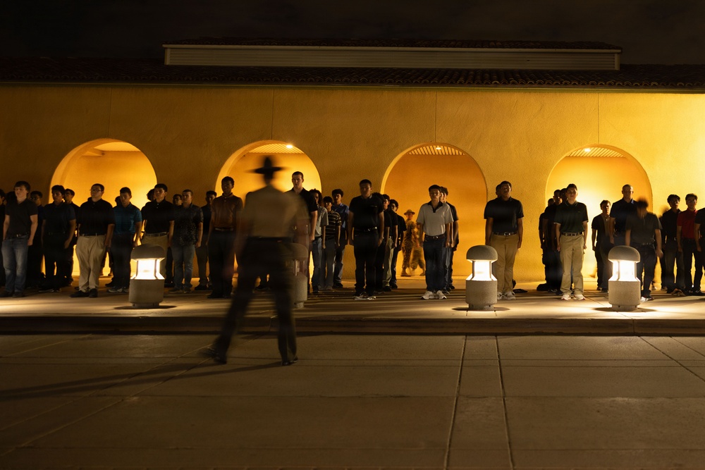 A nighttime outdoor scene showing a large group of young people standing in a line against a yellow stucco wall with illuminated arched alcoves. A single figure wearing a wide-brimmed hat is captured in motion blur in the foreground, suggesting movement or authority. The scene is lit by ground-level lantern-style fixtures and warm lighting from within the arches.