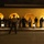 Mike Company Receiving A nighttime outdoor scene showing a large group of young people standing in a line against a yellow stucco wall with illuminated arched alcoves. A single figure wearing a wide-brimmed hat is captured in motion blur in the foreground, suggesting movement or authority. The scene is lit by ground-level lantern-style fixtures and warm lighting from within the arches.