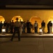 A nighttime outdoor scene showing a large group of young people standing in a line against a yellow stucco wall with illuminated arched alcoves. A single figure wearing a wide-brimmed hat is captured in motion blur in the foreground, suggesting movement or authority. The scene is lit by ground-level lantern-style fixtures and warm lighting from within the arches.