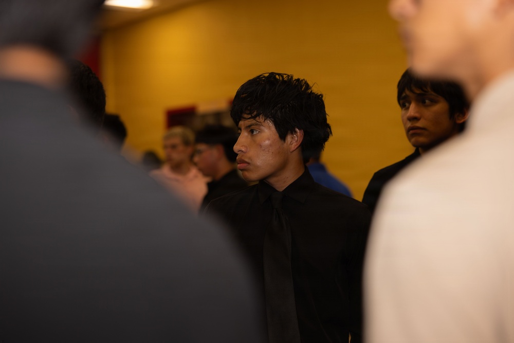 A young man dressed in a black suit and tie stands in the center of the frame, surrounded by a group of people in an indoor setting with a yellow wall in the background. He appears focused or attentive, looking slightly off to the side. The image has a shallow depth of field, with foreground figures blurred.