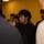 Mike Company Receiving A young man dressed in a black suit and tie stands in the center of the frame, surrounded by a group of people in an indoor setting with a yellow wall in the background. He appears focused or attentive, looking slightly off to the side. The image has a shallow depth of field, with foreground figures blurred.