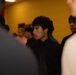 A young man dressed in a black suit and tie stands in the center of the frame, surrounded by a group of people in an indoor setting with a yellow wall in the background. He appears focused or attentive, looking slightly off to the side. The image has a shallow depth of field, with foreground figures blurred.