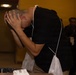 A man in a black t-shirt is bent over a table or surface with his hands clasped over his head in a posture suggesting distress, exhaustion, or emotional overwhelm. He appears to be wearing some kind of lanyard or credential with a mesh pouch. A group of people can be seen standing in the blurred background.