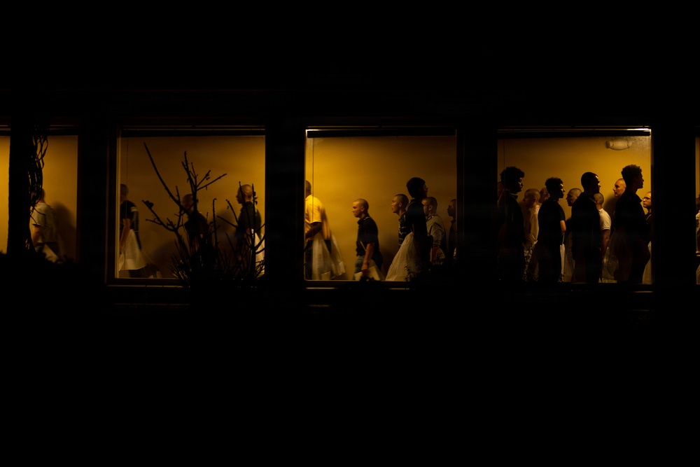 A nighttime exterior photograph looking through illuminated windows into what appears to be an indoor gathering space. Multiple people are visible as silhouettes and partial figures against a warm amber/golden interior light. The dark exterior contrasts sharply with the lit interior, creating a dramatic voyeuristic composition.