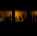 A nighttime exterior photograph looking through illuminated windows into what appears to be an indoor gathering space. Multiple people are visible as silhouettes and partial figures against a warm amber/golden interior light. The dark exterior contrasts sharply with the lit interior, creating a dramatic voyeuristic composition.