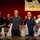 Mike Company Receiving A group of young men with shaved heads stand at attention in rows inside what appears to be a military processing or induction facility. They are holding white mesh bags with white cords/lanyards draped across their chests, suggesting a military intake or processing ceremony. A framed photograph of a military jet aircraft is visible on the red wall in the background.