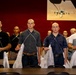 A group of young men with shaved heads stand at attention in rows inside what appears to be a military processing or induction facility. They are holding white mesh bags with white cords/lanyards draped across their chests, suggesting a military intake or processing ceremony. A framed photograph of a military jet aircraft is visible on the red wall in the background.