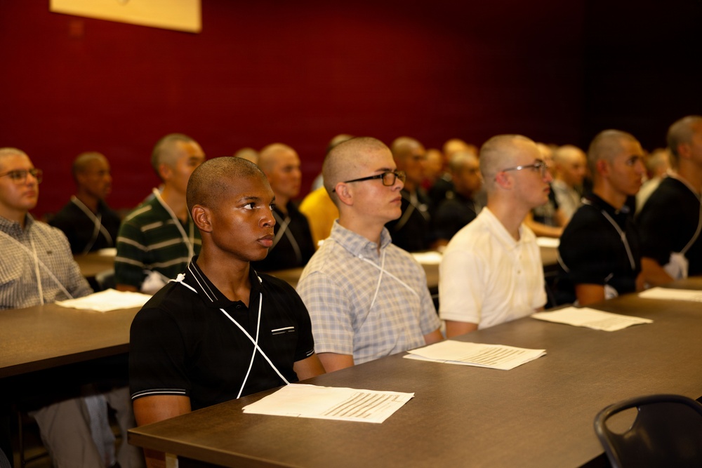 A group of young males with closely cropped or shaved heads are seated at tables in rows, appearing to attend a formal briefing or orientation session. Papers and documents are visible on the tables in front of them. The setting appears to be an institutional or training facility with dark red walls.