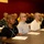 Mike Company Receiving A group of young males with closely cropped or shaved heads are seated at tables in rows, appearing to attend a formal briefing or orientation session. Papers and documents are visible on the tables in front of them. The setting appears to be an institutional or training facility with dark red walls.