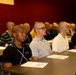 A group of young males with closely cropped or shaved heads are seated at tables in rows, appearing to attend a formal briefing or orientation session. Papers and documents are visible on the tables in front of them. The setting appears to be an institutional or training facility with dark red walls.
