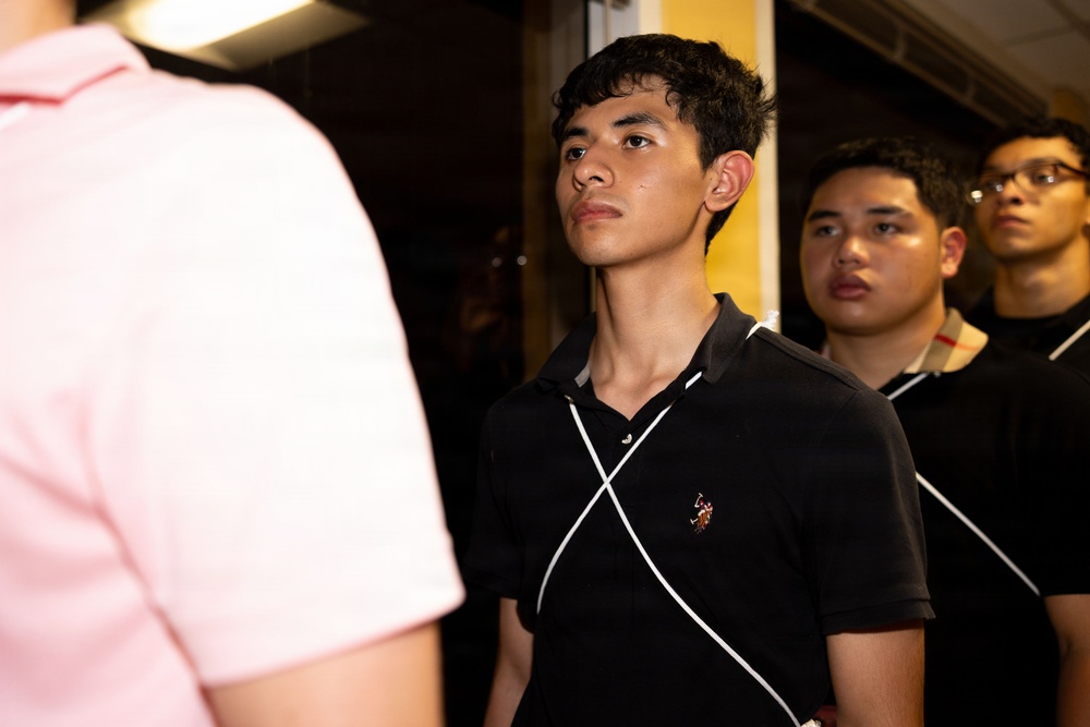 The image shows a group of young men standing together in what appears to be an indoor setting with dim lighting. The central figure, wearing a black polo shirt with white stripe detailing, is looking upward attentively. Two other individuals are partially visible in the background and foreground.