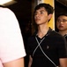 The image shows a group of young men standing together in what appears to be an indoor setting with dim lighting. The central figure, wearing a black polo shirt with white stripe detailing, is looking upward attentively. Two other individuals are partially visible in the background and foreground.