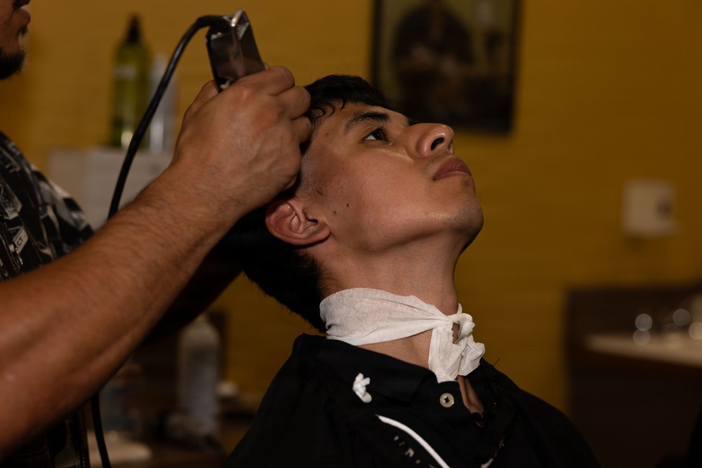 A young man is receiving a haircut at a barbershop, with a barber using electric clippers on the side of his head. The client is seated in a barber chair, wearing a black cape with a white neck strip, and has his head tilted back slightly. The setting is a warmly lit barbershop with yellow walls.