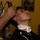 Mike Company Receiving A young man is receiving a haircut at a barbershop, with a barber using electric clippers on the side of his head. The client is seated in a barber chair, wearing a black cape with a white neck strip, and has his head tilted back slightly. The setting is a warmly lit barbershop with yellow walls.