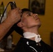 A young man is receiving a haircut at a barbershop, with a barber using electric clippers on the side of his head. The client is seated in a barber chair, wearing a black cape with a white neck strip, and has his head tilted back slightly. The setting is a warmly lit barbershop with yellow walls.