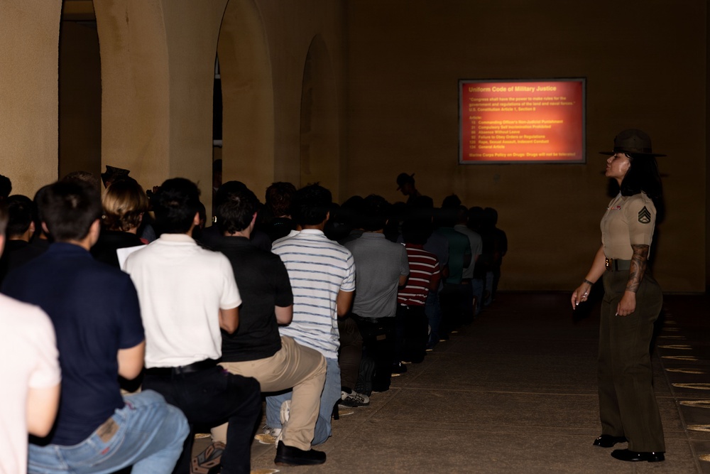 A female Marine Corps drill instructor or staff sergeant stands at attention while overseeing a large group of seated recruits or civilians attending a briefing. A projected presentation titled 'Uniform Code of Military Justice' is displayed on a screen at the front of the room. The setting appears to be a military installation, likely during an orientation or processing event.