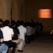 A female Marine Corps drill instructor or staff sergeant stands at attention while overseeing a large group of seated recruits or civilians attending a briefing. A projected presentation titled 'Uniform Code of Military Justice' is displayed on a screen at the front of the room. The setting appears to be a military installation, likely during an orientation or processing event.