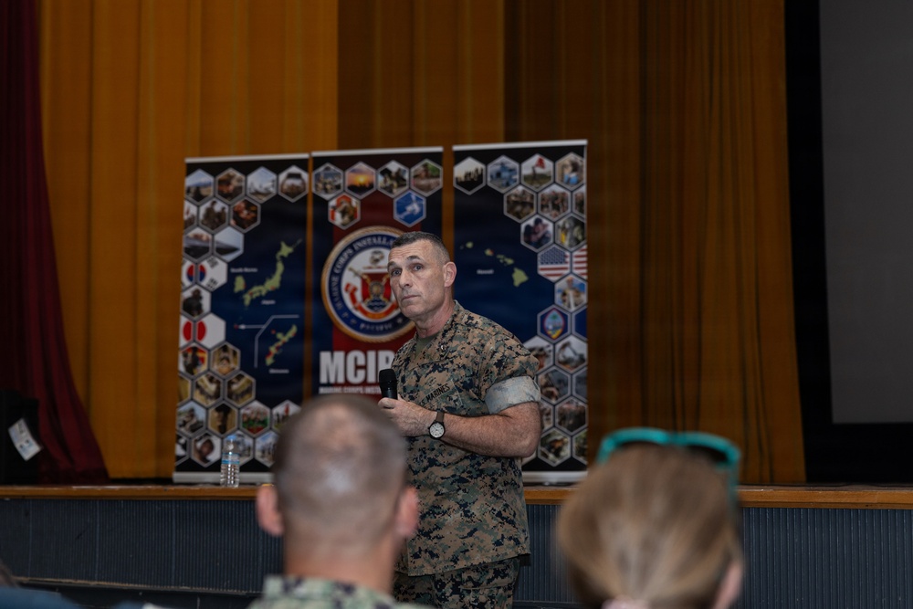 A U.S. Marine Corps officer in digital camouflage (MARPAT) uniform is addressing an audience while holding a microphone, standing in front of MCIPAC (Marine Corps Installations Pacific) banner displays. The setting appears to be an auditorium or theater with a stage, curtains, and a projection screen. Several audience members are visible in the foreground, including what appears to be military and civilian personnel.