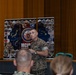 A U.S. Marine Corps officer in digital camouflage (MARPAT) uniform is addressing an audience while holding a microphone, standing in front of MCIPAC (Marine Corps Installations Pacific) banner displays. The setting appears to be an auditorium or theater with a stage, curtains, and a projection screen. Several audience members are visible in the foreground, including what appears to be military and civilian personnel.