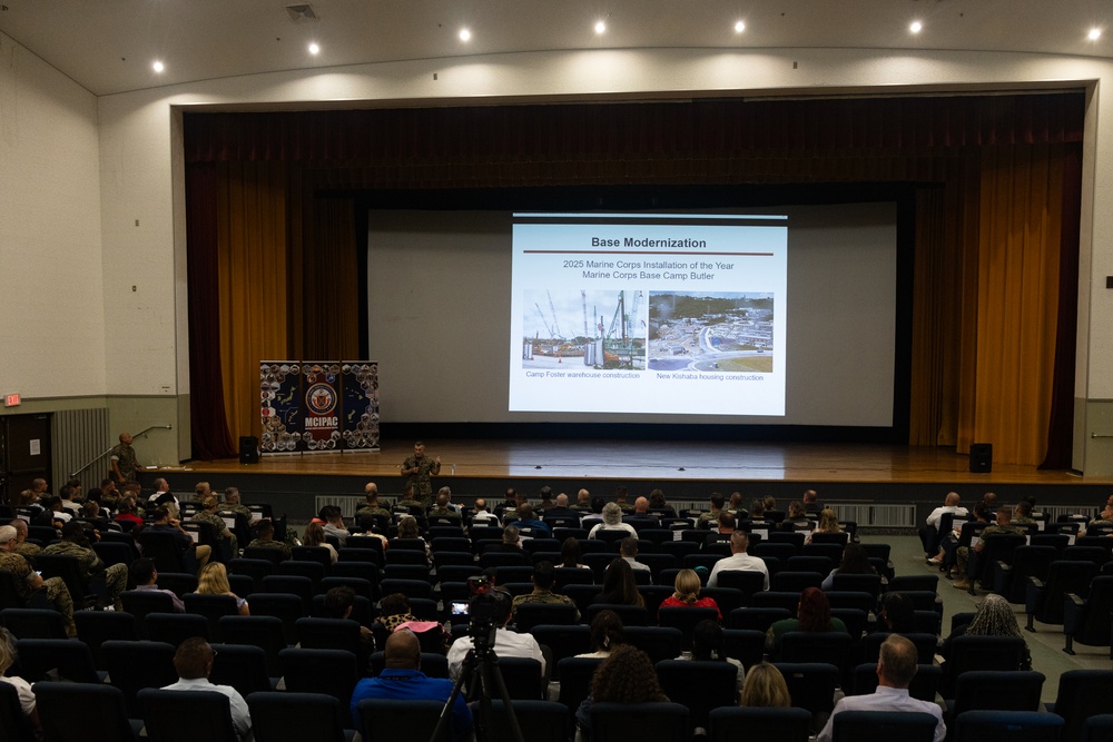 A military briefing or town hall is taking place in an auditorium, with an audience of military personnel and civilians seated in rows facing a stage. A presenter stands on stage next to a projected slideshow titled 'Base Modernization' referencing Marine Corps Base Camp Butler and the '2025 Marine Corps Installation of the Year.' The presentation includes construction photos labeled 'Camp Foster warehouse construction' and 'New Kishaba housing construction.'