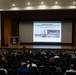 A military briefing or town hall is taking place in an auditorium, with an audience of military personnel and civilians seated in rows facing a stage. A presenter stands on stage next to a projected slideshow titled 'Base Modernization' referencing Marine Corps Base Camp Butler and the '2025 Marine Corps Installation of the Year.' The presentation includes construction photos labeled 'Camp Foster warehouse construction' and 'New Kishaba housing construction.'