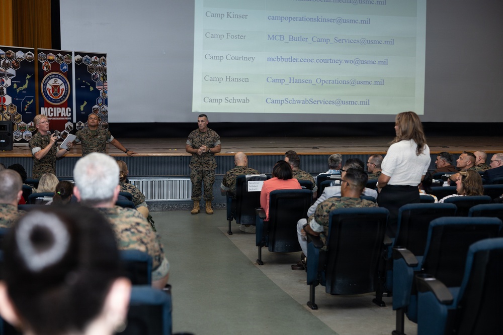 A military briefing or town hall meeting is taking place in an auditorium-style venue under the MCIPAC (Marine Corps Installations Pacific) banner. Three uniformed Marines are presenting at the front of the room to an audience of both military personnel and civilians. A projection screen displays contact email addresses for various Marine Corps camps including Camp Kinser, Camp Foster, Camp Courtney, Camp Hansen, and Camp Schwab.
