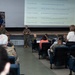 A military briefing or town hall meeting is taking place in an auditorium-style venue under the MCIPAC (Marine Corps Installations Pacific) banner. Three uniformed Marines are presenting at the front of the room to an audience of both military personnel and civilians. A projection screen displays contact email addresses for various Marine Corps camps including Camp Kinser, Camp Foster, Camp Courtney, Camp Hansen, and Camp Schwab.