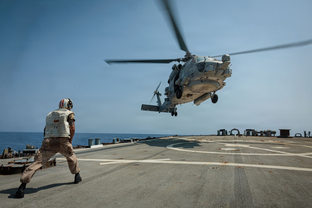 A military helicopter, appearing to be an MH-60 Seahawk, is hovering or landing on the flight deck of a naval vessel at sea. A flight deck crew member wearing a white helmet and safety vest is positioned on the deck, likely guiding the aircraft. The scene takes place under clear skies over open ocean.