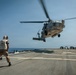 A military helicopter, appearing to be an MH-60 Seahawk, is hovering or landing on the flight deck of a naval vessel at sea. A flight deck crew member wearing a white helmet and safety vest is positioned on the deck, likely guiding the aircraft. The scene takes place under clear skies over open ocean.