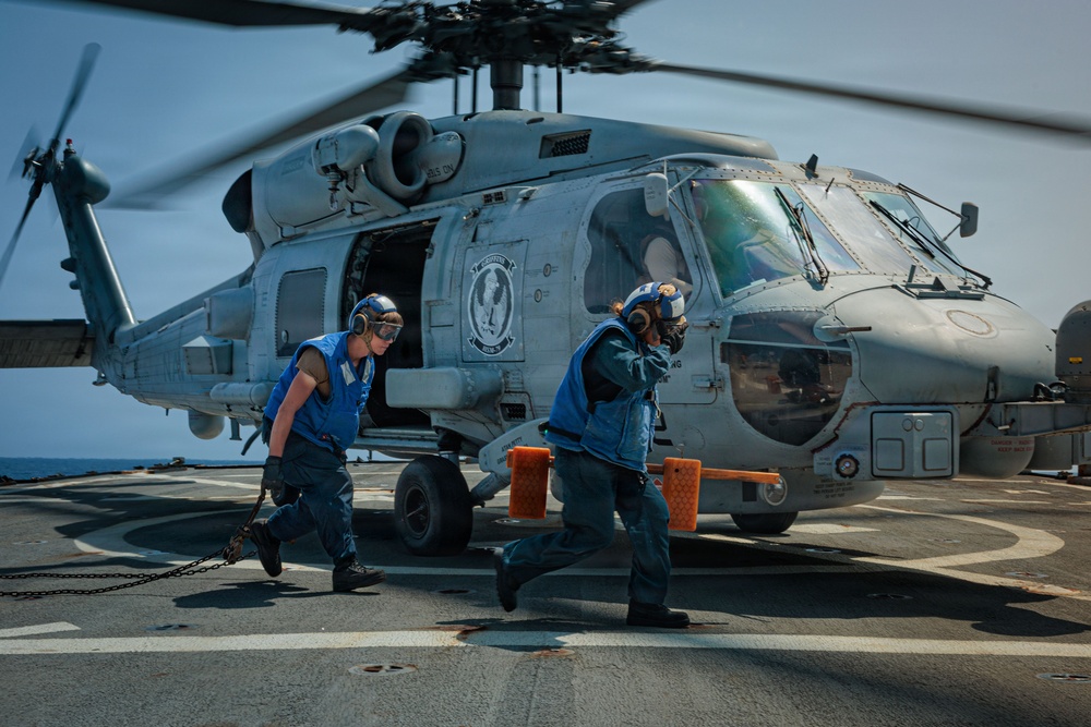 Two naval deck crew members in blue vests and hearing protection are running across a ship's flight deck near a military helicopter, likely an MH-60 Seahawk. The helicopter's rotors are spinning, indicating active flight operations on what appears to be a U.S. Navy vessel. The crew members appear to be removing wheel chocks or performing pre/post-flight deck operations.