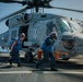 Two naval deck crew members in blue vests and hearing protection are running across a ship's flight deck near a military helicopter, likely an MH-60 Seahawk. The helicopter's rotors are spinning, indicating active flight operations on what appears to be a U.S. Navy vessel. The crew members appear to be removing wheel chocks or performing pre/post-flight deck operations.
