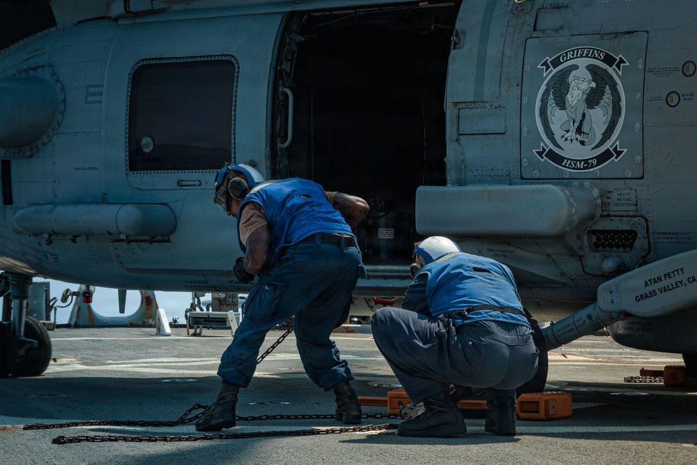Two U.S. Navy personnel in blue uniforms and white helmets are performing maintenance or chocking/chaining operations on a military helicopter aboard a ship's flight deck. The helicopter bears the insignia of HSM-79 'Griffins,' a helicopter maritime strike squadron. The scene takes place on what appears to be a naval vessel at sea under bright daylight conditions.