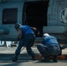 Two U.S. Navy personnel in blue uniforms and white helmets are performing maintenance or chocking/chaining operations on a military helicopter aboard a ship's flight deck. The helicopter bears the insignia of HSM-79 'Griffins,' a helicopter maritime strike squadron. The scene takes place on what appears to be a naval vessel at sea under bright daylight conditions.